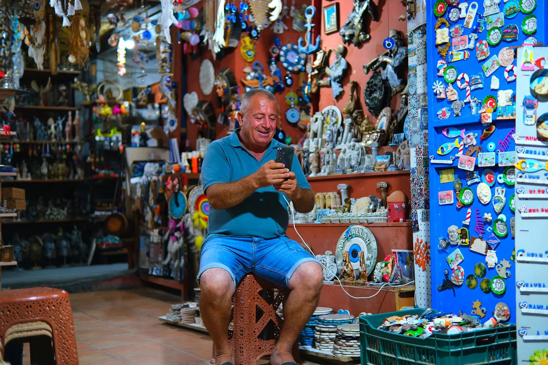 Lojista sentado dentro de uma loja de lembranças, sorrindo enquanto olha o celular