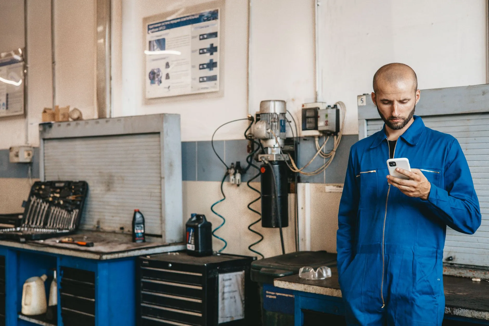 Mecânico de macacão azul olhando o celular, em pé dentro de uma oficina, com bancada de ferramentas ao fundo